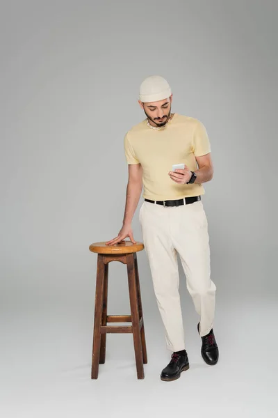 Повна довжина модного гомосексуального чоловіка в капелюсі за допомогою смартфона біля стільця на сірому фоні — стокове фото