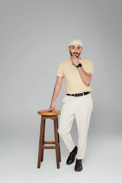 Elegante uomo omosessuale in cappello e orologio intelligente in piedi vicino sedia su sfondo grigio — Foto stock
