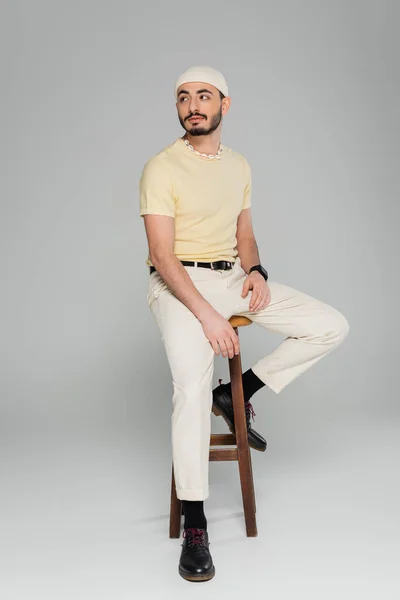 Joven hombre homosexual en sombrero beige mirando hacia otro lado mientras está sentado en la silla sobre fondo gris - foto de stock