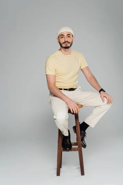 Lunghezza completa dell'uomo omosessuale alla moda seduto sulla sedia su sfondo grigio — Foto stock