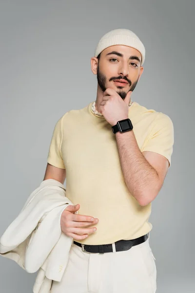 Ritratto di uomo gay elegante in cappello con giacca beige e mento toccante isolato sul grigio — Foto stock