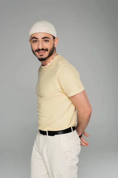 Retrato de hombre homosexual de moda en sombrero y collar de concha sonriendo a la cámara aislada en gris - foto de stock