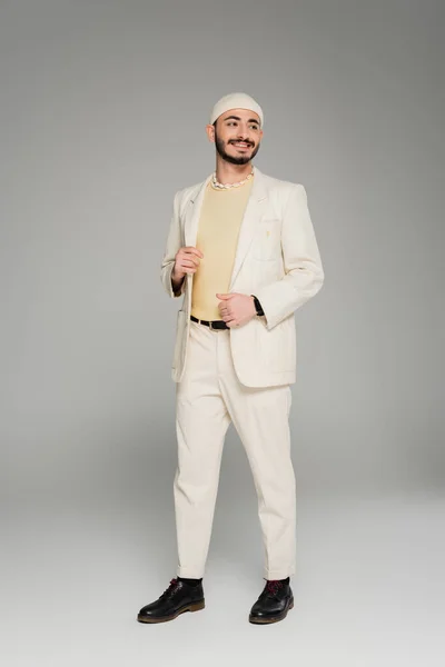 Cheerful homosexual man in beige suit and hat touching jacket on grey background — Stock Photo