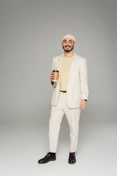 Alegre gay hombre en traje con lgbt bandera en mejilla celebración papel taza en gris fondo - foto de stock