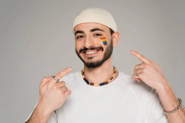 Smiling homosexual man in hat pointing at lgbt flag on cheek isolated on grey — Stock Photo