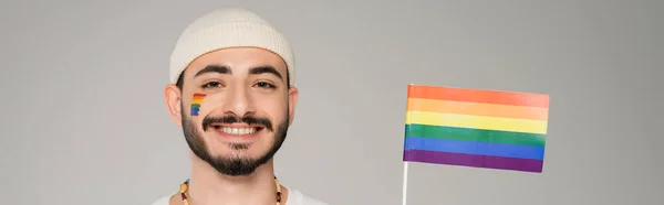 Веселый гомосексуалист в шляпе смотрит в камеру рядом с флагом lgbt, изолированным на сером, баннер — стоковое фото