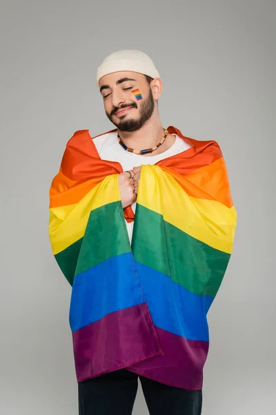 Gioioso gay uomo holding lgbt bandiera e chiusura occhi isolato su grigio — Foto stock