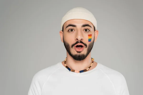 Schockierter schwuler Mann mit Hut und lgbt-Fahne auf der Wange blickt isoliert in die Kamera auf grau — Stockfoto