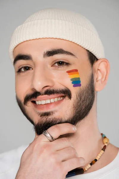 Retrato de sorrindo gay homem no chapéu com lgbt bandeira no rosto tocando queixo isolado no cinza — Fotografia de Stock