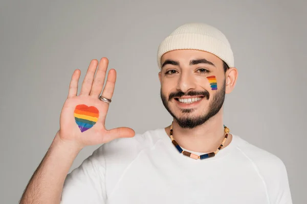 Улыбающийся гомосексуалист с вывеской сердца от флага lgbt на руке, изолированный на сером — стоковое фото