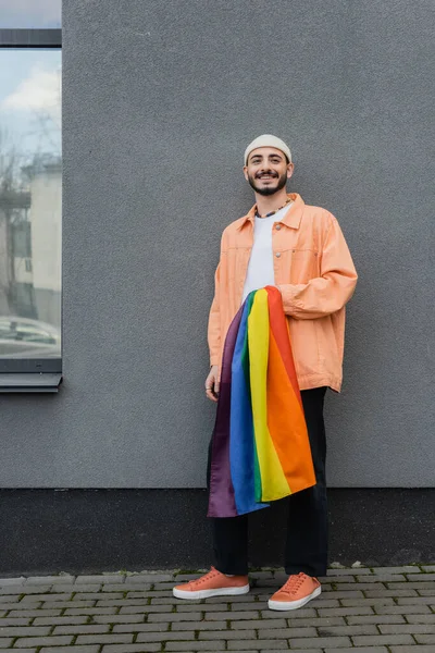 Smiling gay man holding lgbt flag near building outdoors — Stock Photo