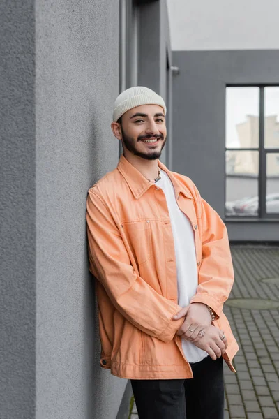 Stylischer schwuler Mann mit Hut und Jacke lächelt in die Kamera in der Nähe eines Gebäudes an der Stadtstraße — Stockfoto