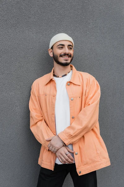 Trendy e positivo gay uomo in cappello e giacca in piedi vicino al muro sulla strada urbana — Foto stock