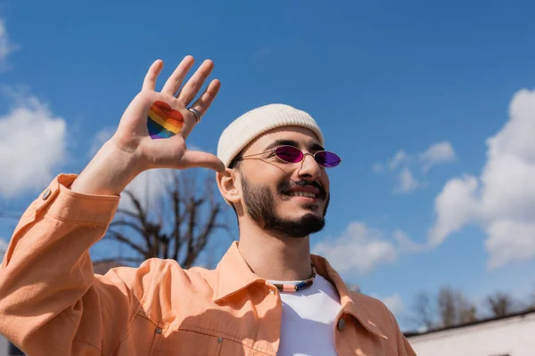 Alegre gay homem no sol óculos com lgbt bandeira no coração forma no mão pé ao ar livre — Fotografia de Stock