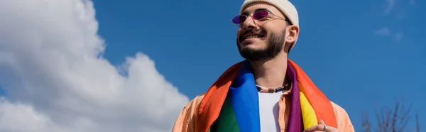Low angle view of carefree gay man in sunglasses with lgbt flag standing on street, banner — Stock Photo