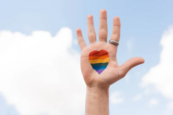 Vista recortada del hombre gay con bandera lgbt de la señal del corazón y el cielo en el fondo, Día Internacional contra la homofobia - foto de stock