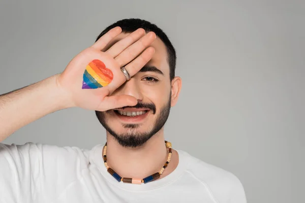 Улыбающийся гей, закрывающий глаза с флагом lgbt на руке, изолированный в серый день, Международный день борьбы с гомофобией — стоковое фото