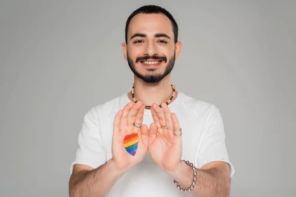 Joven hombre gay sonriente con bandera lgbt en la mano mirando a la cámara aislada en gris, Día internacional contra la homofobia - foto de stock
