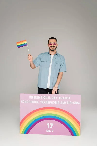 Allegro gay uomo tenendo lgbt bandiera vicino cartello con internazionale giornata contro omofobia, transfobia e bifobia lettering su sfondo grigio — Foto stock