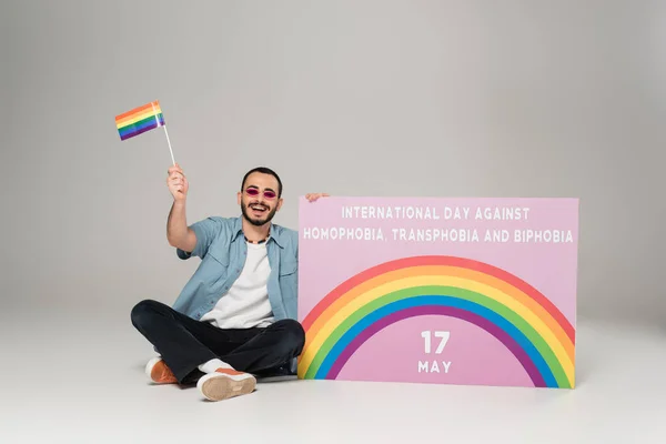Позитивный гей, держащий флаг lgbt возле плаката с Международным днем борьбы с гомофобией, трансфобией и бифобией на сером — стоковое фото