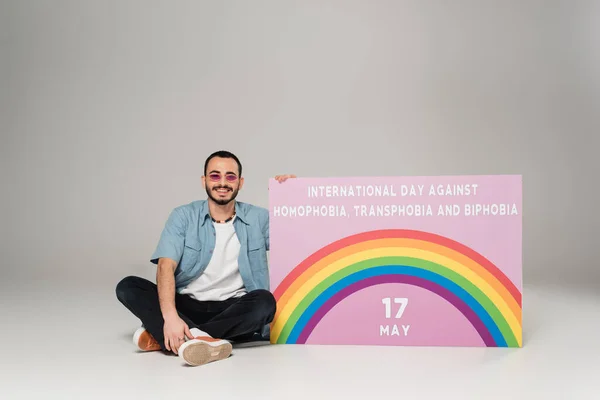Cheerful gay man near placard with International Day Against Homophobia, Transphobia and Biphobia lettering on grey — Stock Photo