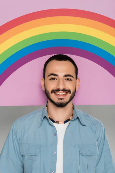 Портрет улыбающегося гея, смотрящего в камеру рядом с плакатом с флагом lgbt, изолированным на сером — стоковое фото