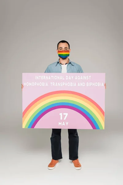 Gay uomo in medico maschera con lgbt bandiera holding cartello con internazionale giornata contro omofobia, transfobia e bifobia lettering su grigio — Foto stock