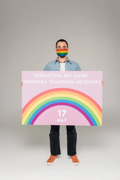 Gay homem no médico máscara com lgbt bandeira segurando cartaz com internacional dia contra homofobia Transphobia e bifobia lettering no cinza — Fotografia de Stock