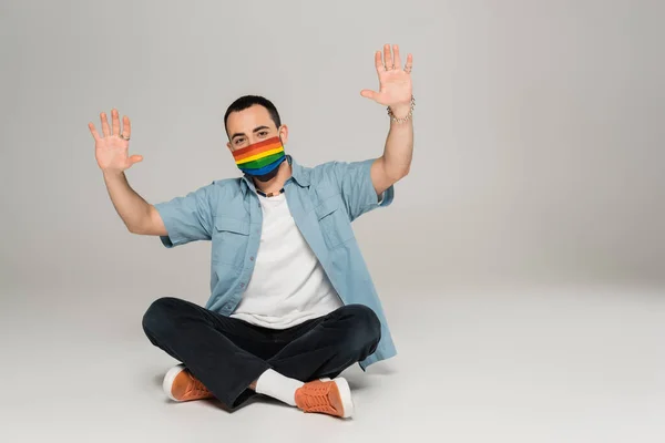 Брюнетка гей мужчина в медицинской маске в цветах флага lgbt размахивая руками на сером фоне — стоковое фото