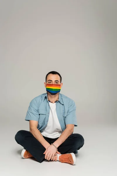 Young homosexual man in medical mask with lgbt flag sitting on grey background — Stock Photo