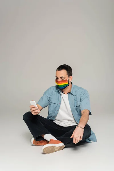 Гомосексуальна людина в медичній масці з кольорами прапора lgbt, використовуючи смартфон на сірому фоні — стокове фото