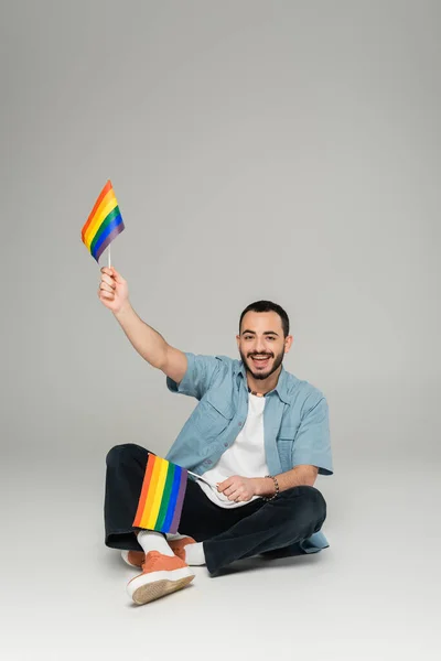 Усміхнений гей чоловік тримає lgbt прапори, сидячи на сірому фоні — стокове фото