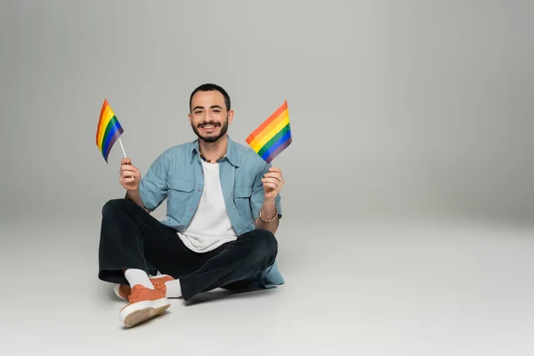 Alegre hombre homosexual sosteniendo banderas lgbt y mirando a la cámara mientras está sentado sobre fondo gris - foto de stock