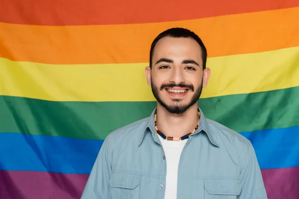 Alegre gay hombre mirando a cámara cerca lgbt bandera en fondo, internacional día contra homofobia - foto de stock