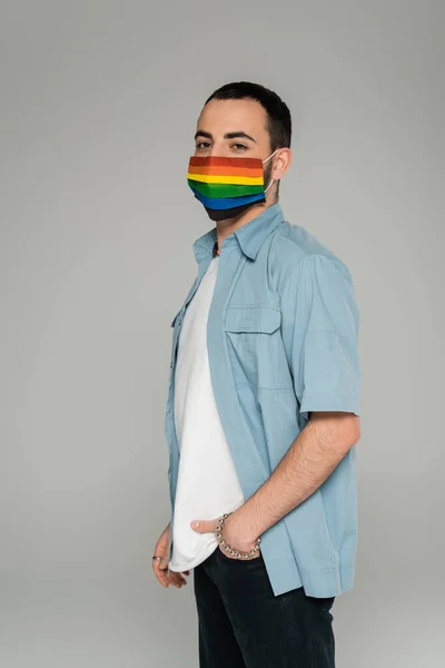 Bruna gay uomo in medico maschera con lgbt bandiera guardando fotocamera isolato su grigio — Foto stock