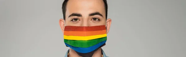 Giovane bruna gay uomo in medico maschera con lgbt bandiera guardando fotocamera isolato su grigio banner — Foto stock