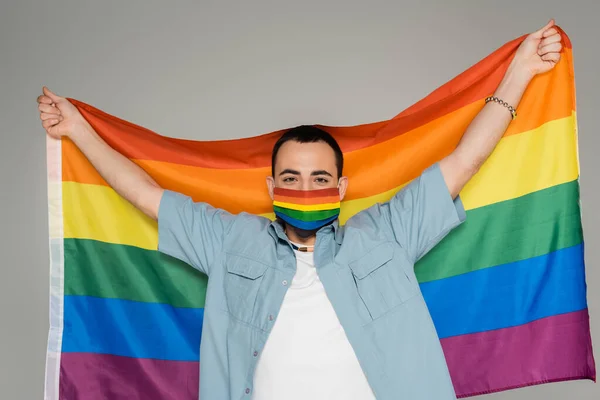 Молодой гей в медицинской маске держит флаг lgbt изолированным на сером, Международный день борьбы с гомофобией — стоковое фото