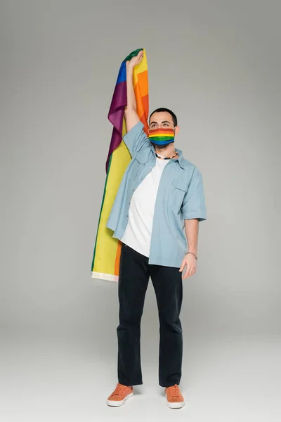 Повна довжина молодого гея в медичній масці, що тримає прапор lgbt на сірому фоні — стокове фото
