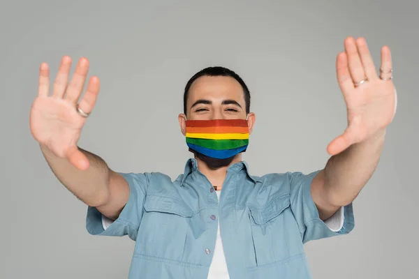 Morena gay homem no médico máscara no cores de lgbt bandeira estendendo as mãos isolado no cinza — Fotografia de Stock