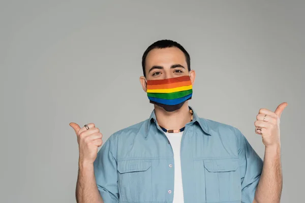 Brünetten homosexuellen Mann mit medizinischer Maske in lgbt Farben zeigt wie Geste isoliert auf grau, Internationaler Tag gegen Homophobie — Stockfoto