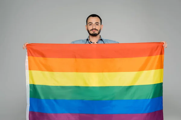Barbuto omosessuale uomo tenendo lgbt bandiera e guardando fotocamera isolato su grigio — Foto stock
