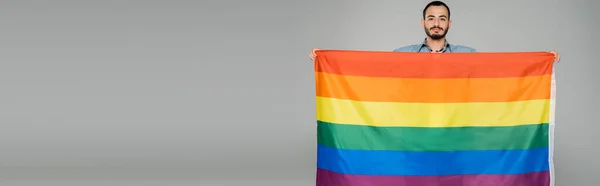 Bruna gay uomo tenendo lgbt bandiera e guardando fotocamera isolato su grigio banner — Foto stock
