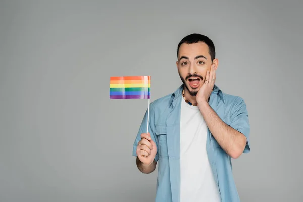Изумленный гомосексуалист трогает лицо и держит флаг lgbt изолированным на сером, Международный день борьбы с гомофобией — стоковое фото