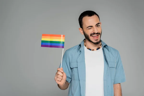 Hombre homosexual despreocupado sosteniendo bandera lgbt y guiñando un ojo a la cámara aislado en gris, Día Internacional contra la Homofobia - foto de stock