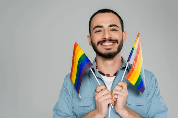 Feliz hombre homosexual sosteniendo banderas lgbt y mirando a la cámara aislada en gris - foto de stock