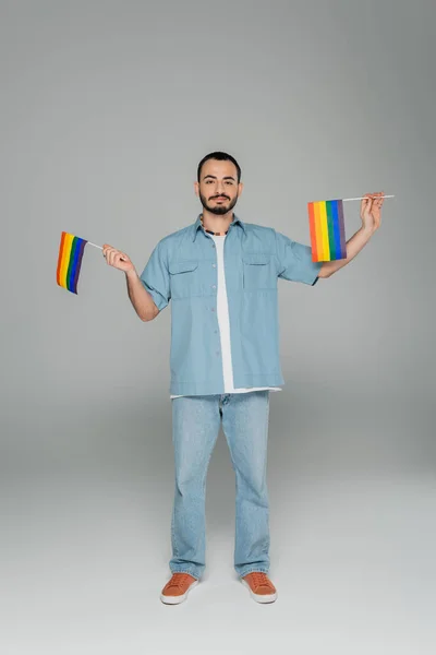 Full length of young gay man holding lgbt flags while standing on grey background, International Day Against Homophobia — Stock Photo