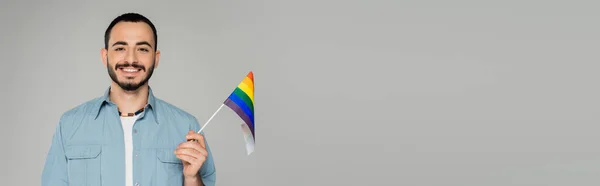 Bruna gay uomo sorridente a fotocamera e tenendo lgbt bandiera isolato su grigio banner — Foto stock