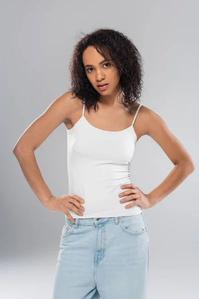 Esbelta mujer afroamericana en camiseta blanca con las manos en las caderas sobre fondo gris - foto de stock
