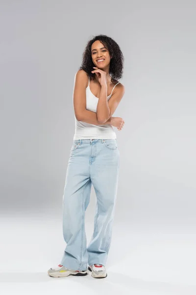 Longitud completa de la alegre mujer afroamericana en camiseta blanca y pantalones vaqueros azules sobre fondo gris - foto de stock