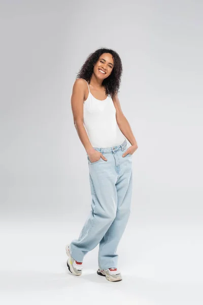 Pleine longueur de souriante femme afro-américaine posant avec les mains dans des poches de jeans bleus sur fond gris — Photo de stock